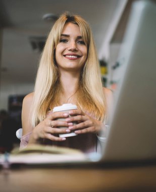 Uzun sarı saçlı, kolsuz elbiseli, kahve dükkanında oturan, elinde kahve fincanı olan ve kameraya gülümseyen bir kadın.