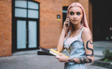 Dövmeli, uzun pembe saçlı, kot elbiseli, elinde sarı kitapla cep telefonuyla konuşan şık bir kadın.