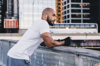 Spor giyim giyim giymiş kel sakallı bir zenci şehir merkezindeki ahşap iskelede korkuluklara yaslanıp kulaklıkla müzik dinliyor.
