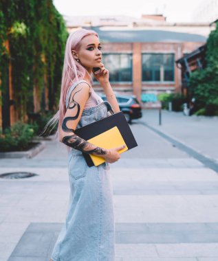 Dövmeli kollu, pembe saçlı, not defteri taşıyan ve sokakta yürürken akıllı telefon konuşması yapan şık bir genç kadın.