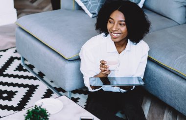 Kahvesini ve dijital dokunmatik defterini elinde tutarak gülen koyu tenli hippi blogcu modern bir dairede şık bir iç dekorasyonla oturuyor. Neşeli, düşünceli, Afro-Amerikan genç kadın.