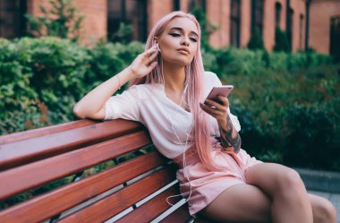 Beyaz gömlekli, pembe şortlu, uzun pembe saçlı, akıllı telefondan müzik dinleyen, tuğla binanın karşısındaki bankta oturan genç bir bayan.