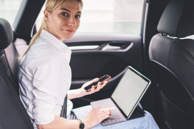 Gülümseyen kadın yönetici emniyet kemeriyle arabanın arka koltuğuna bağlanmış, akıllı telefon tutuyor ve kameraya bakarken tablet klavyeye yazı yazıyor.