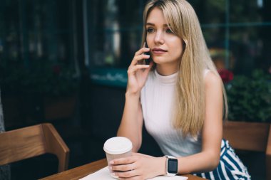 Sarışın, kolsuz balıkçı yaka giyen genç bir kadın kahve dükkanının önündeki masada oturuyor ve elinde bir fincan kahveyle telefonla konuşuyor.