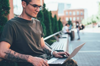 Gözlüklü ve sıradan kıyafetleri olan genç bir adamın ara sokakta bankta otururken dizüstü bilgisayarda yazışının yan görüntüsü.