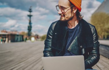 Moda giyinmiş hipster bir adamın portresi dizüstü bilgisayarla dışarıda oturup 4G internet bağlantısıyla tatmin olan başarılı erkek serbest yazar tanıtım alanında güneşli bir günün tadını çıkarıyor.