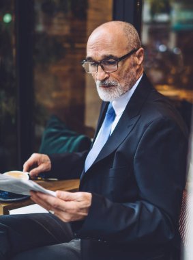 Sakallı, kahve bardaklı, takım elbiseli, beyaz duvarın yanında oturan ve gazete okuyan yaşlı adamın yan görüntüsü.