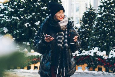 Noel ağaçlarının yanında duran ve karlı caddeye bakan, kağıt bardak ve telefonla şık kış kıyafetleri içindeki alımlı genç bayan.
