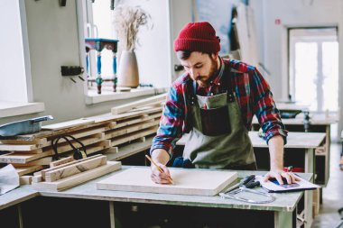 Ekose gömlekli, kırmızı şapkalı ve gri önlüklü düşünceli, çalışkan marangozun yüksek açısı. Masada dikilirken kalemle konsantre oluyor ve çiziyor. 