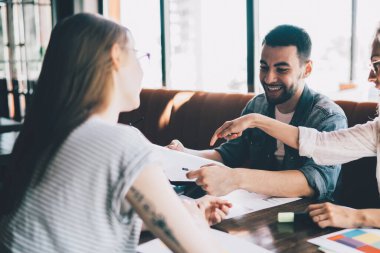 Bir grup modern genç kadın ve erkek masada kağıt ve çalışmayla birlikte otururken aynı zamanda takım halinde yeni bir proje üzerinde çalışıyorlar.