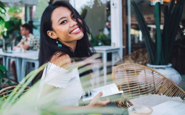 Beyaz omuzlu, kitap okuyan, terasta bir fincan kahveyle oturmuş gülümseyen Asyalı güzel bir kadının yan görüntüsü.