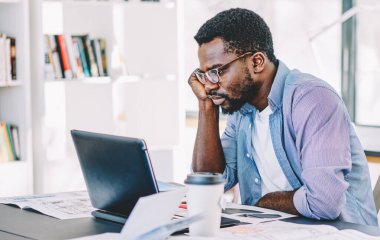 Gözlüklü, tişörtlü, dizüstü bilgisayarlı bir Afro-Amerikan adam gözlüklü, not defterleriyle dolu bir masada oturuyor.