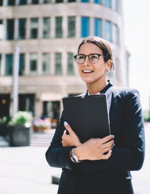 Cilalı tırnakları parlak genç bir bayan ajan iş merkezinin yanında duruyor göğsüne yakın bir pano tutuyor ve yüzünde gülümsemeyle yüzünü çeviriyor.