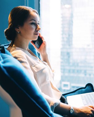 Zarif elbiseli, düşünceli yetişkin bir kadının telefon görüşmesi yapıp koltukta otururken gülümsemesinin yan görüntüsü. 