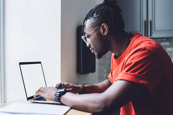 Side view of modern African American man in glasses sitting at counter with notepad and typing on laptop with empty screen