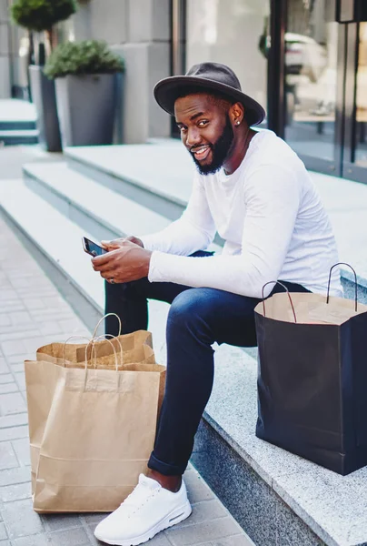 Portrait of afro american guy waiting for feedback from web store service sitting on stairs waiting for purchase via smartphone, positive dark skinned man in stylish hat resting after shopping time