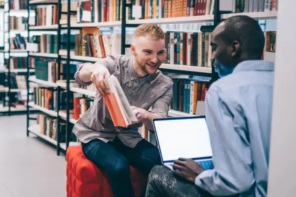 Cheerful young guy showing textbook to black classmate with laptop while sitting near woman and bookshelves and studying in library
