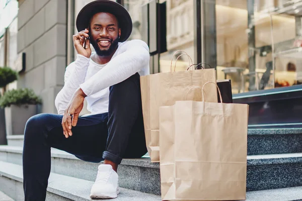 Happy afro american male buyer in trendy wear calling to friend for communicate about successful shopping, cheerful dark skinned guy having positive mobile conversation sitting on stairs with bags