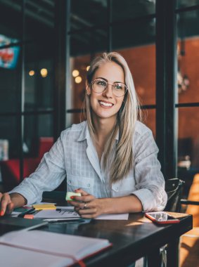 Kafedeki masada dizüstü bilgisayarı ve ofis malzemeleriyle oturmuş düşünceli bir şekilde yüzünü çevirmiş mutlu bir şekilde gülümseyen güzel sarışın.