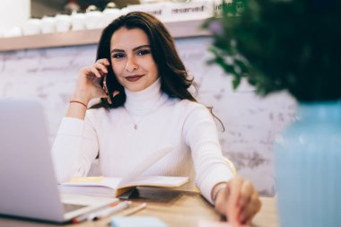 Beyaz kazaklı, esmer, neşeli ve zeki bir bayan cep telefonuyla konuşuyor. İş yerinde dizüstü bilgisayarında renkli etiketlerle oturuyor.