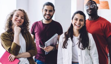 Kağıtları ve not defterleriyle ışık salonunda bir arada duran ve kameraya neşeyle gülümseyen bir grup mutlu ve çok ırklı genç öğrenci.