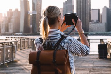 Amerikan kentsel ortamında duran gezici sırt çantalı şık bayan turistin arka planda görüntüsü ve Manhattan şehir simgesinin resimlerine retro anlık kamera kullanarak tıklamak, fotoğrafçılık hobisi