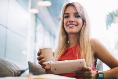 Bir fincan içkisi olan mutlu genç bir bayan kahve dükkanında oturmuş kameraya bakıp gülümsüyor.