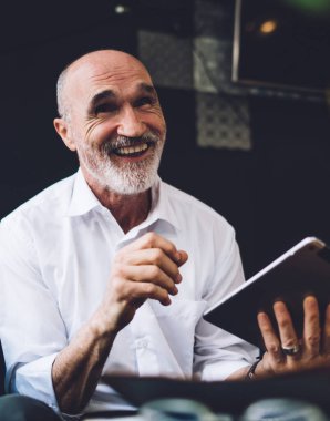 Beyaz tişörtlü, gri sakallı, elinde tabletle oturma odasında oturan mutlu ve kel bir adam.