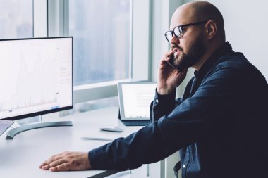 Ofiste cep telefonuyla konuşurken bilgisayar ve dizüstü bilgisayarla çalışan gözlüklü yetişkin kel bir adam.