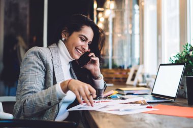 Mutlu bayanın iş arkadaşıyla telefonda konuşurken bilgisayarla kafede oturarak evrakları analiz ederken yan görüntüsü.