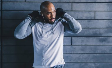 Beyaz kulaklıklı, siyah sırt çantalı, kaputu tamir ederken duvara yaslanan ve fotokopi çeken gri spor giysili kel bir sporcu.