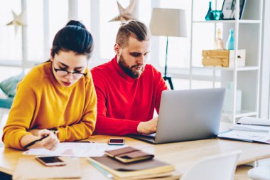 Kafkasyalı öğrenciler, evdeki iç mekanda birlikte oturarak projede işbirliği yapıyorlar. Meslektaşlar, kadın belgelerden muhasebe kayıtlarını incelerken erkek daktilo yazmayı planlamaya odaklanmış durumdalar.
