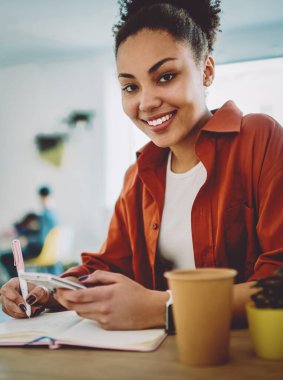 Mutlu kız öğrencinin portresi üniversite kafeteryasında gençlerin otostop çekerken eğlenmesi, elinde cep telefonu ve ders kitabı olan pozitif hippi kız.