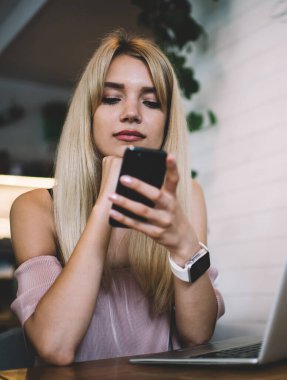Akıllı saat takan çağdaş kız öğrenci laptopun önündeki kafede otururken cep telefonundan mesajları kontrol ediyor.