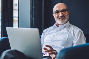 Gözlüklü, resmi giyinmiş, laptopun önündeki kafede oturan ve kameraya bakıp yazarken bacak bacak bacak üstüne atan neşeli yaşlı girişimci.