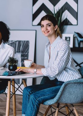 Elinde kahve fincanı olan, kameraya gülümseyen ve modern dairede şık iç dekorasyonu olan Afro-Amerikan arkadaşıyla aynı masada oturan beyaz bir kadının portresi.