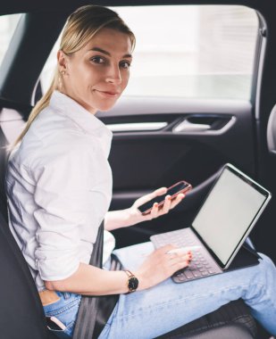 Gülümseyen kadın yönetici emniyet kemeriyle arabanın arka koltuğuna bağlanmış, akıllı telefon tutuyor ve kameraya bakarken tablet klavyeye yazı yazıyor.