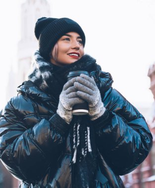 Büyük beden ceketli mutlu genç bir kadın tarihi binaların yanında dinleniyor. Plastik bardakta sıcak bir kahve ve bulanık bir arka plan.