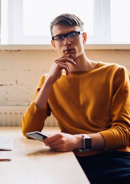 Gözlüklü ciddi bir hippi öğrencinin modern ofiste bilgisayarla otururken kameraya baktığı portre. Elinde cep telefonu tutan genç bir adam.