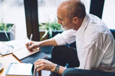 Beyaz tişörtlü, sakallı, tabletini karıştıran yaşlı bir adamın yan görüntüsü. Not defterine yazarken ve kafede oturmuş çay içerken.