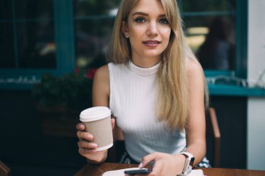 Bir fincan kahve içerken ve kahve masasında otururken cep telefonu kullanan uzun saçlı genç bir kadın.