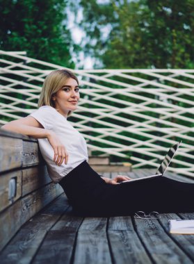 Sıradan kıyafetli genç bir kadının kitap ve dizüstü bilgisayarla ahşap tribünde oturup kameraya bakarken yan görüntüsü.