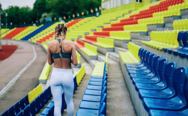 Modern spor giysileri içinde ince bir kadın ve örgülü saçlarıyla stadyumdaki renkli tribünlerin arasında yürüyor.