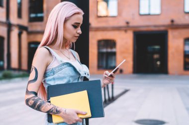Pembe saçlı, dövmeli, elinde defterler olan ve akıllı telefonları gezen şık genç bir kadın.
