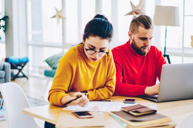 Kafkasyalı öğrenciler, evdeki iç mekanda birlikte oturarak projede işbirliği yapıyorlar. Meslektaşlar, kadın belgelerden muhasebe kayıtlarını incelerken erkek daktilo yazmayı planlamaya odaklanmış durumdalar.