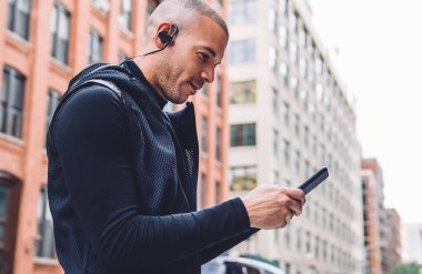 Siyah spor ceket giyen ve kulaklık takan etnik bir adamın şehrin caddesinde telefon kullanarak uygulamadan müzik seçerken yan görüntüsü