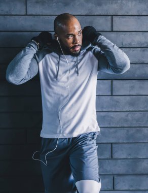 Beyaz kulaklıklı, siyah sırt çantalı, kaputu tamir ederken duvara yaslanan ve fotokopi çeken gri spor giysili kel bir sporcu.