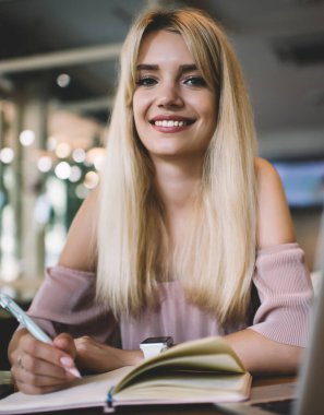 Açık pembe elbise giyen, gülümseyen ve kameraya bakan genç bir sarışın. Not defterine yazarken ve laptopla kafede otururken.