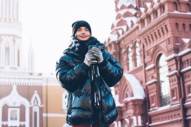 Koyu renk kışlık ceketli tatmin olmuş genç bir kadın açık gökyüzünde plastik bardakta sıcak kahveyle tarihi binaların yanında duruyor.