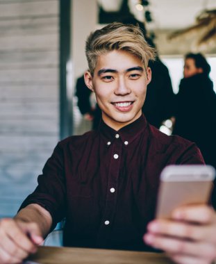 Neşeli Çinli yenilikçi adam sosyal ağlarda akıllı telefondan komik videolar izliyor modern mekanda oturuyor. Japon blogcu kendi web sitesinden 4G internet kullanarak yayın indiriyor.